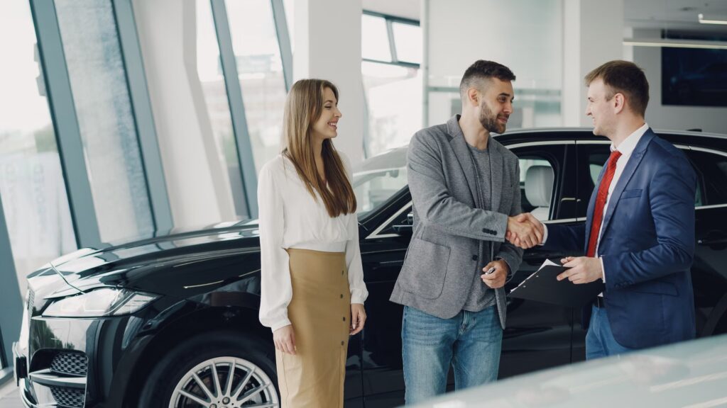 A car dealer shaking hands with a couple in front of a vehicle in a showroom.