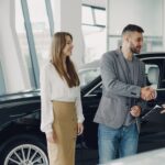 A car dealer shaking hands with a couple in front of a vehicle in a showroom.